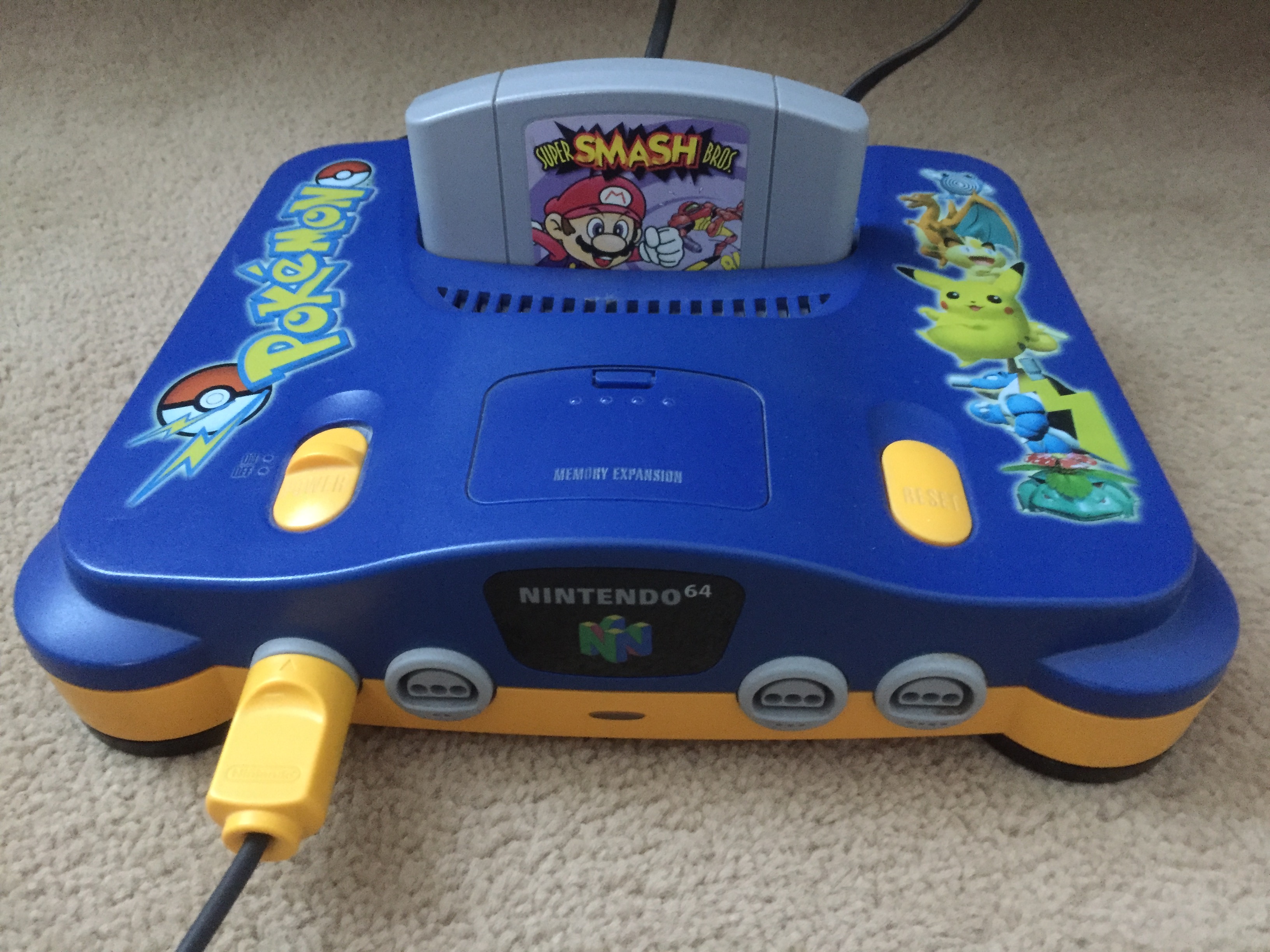 A Nintendo 64 console with the Super Msash Bros cartridge plugged in. The console is a Pokémon Stadium special edition. The upper half is blue, the bottom yellow. The Pokémon logo is on the left on the top of the console and several 3D renderings of Pokémon on the right.